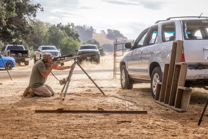 Kneeling rifle on bipod beside a shot-up SUV