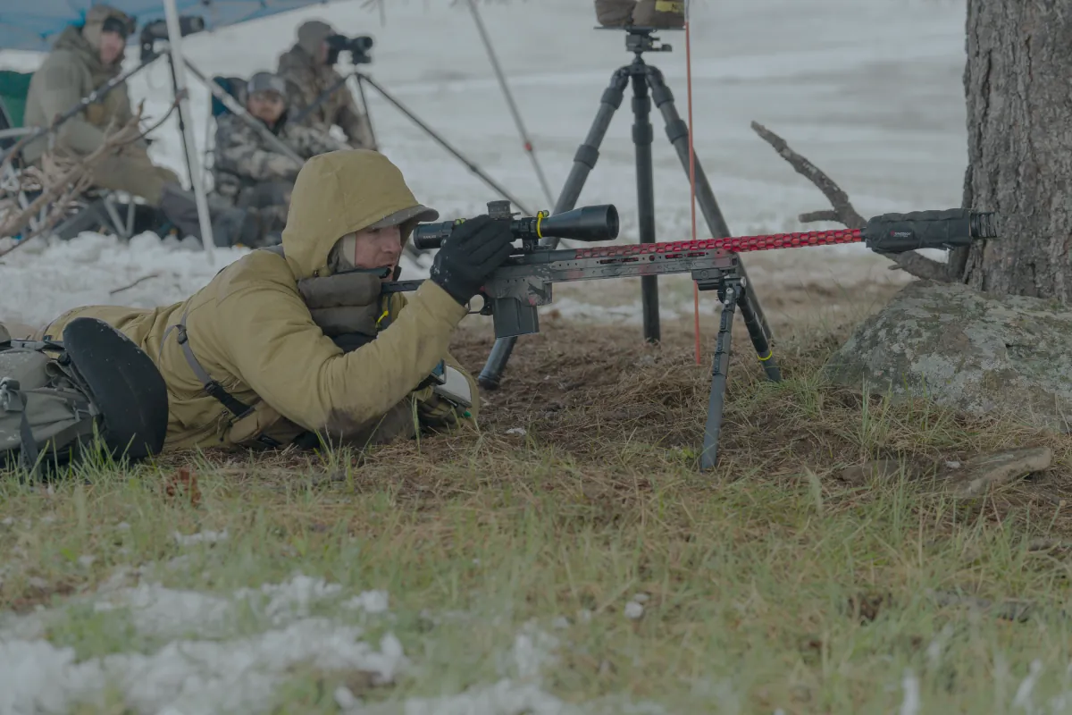 A Cadre shooter prone in the snow with spotters glassing behind at a cold-weather match