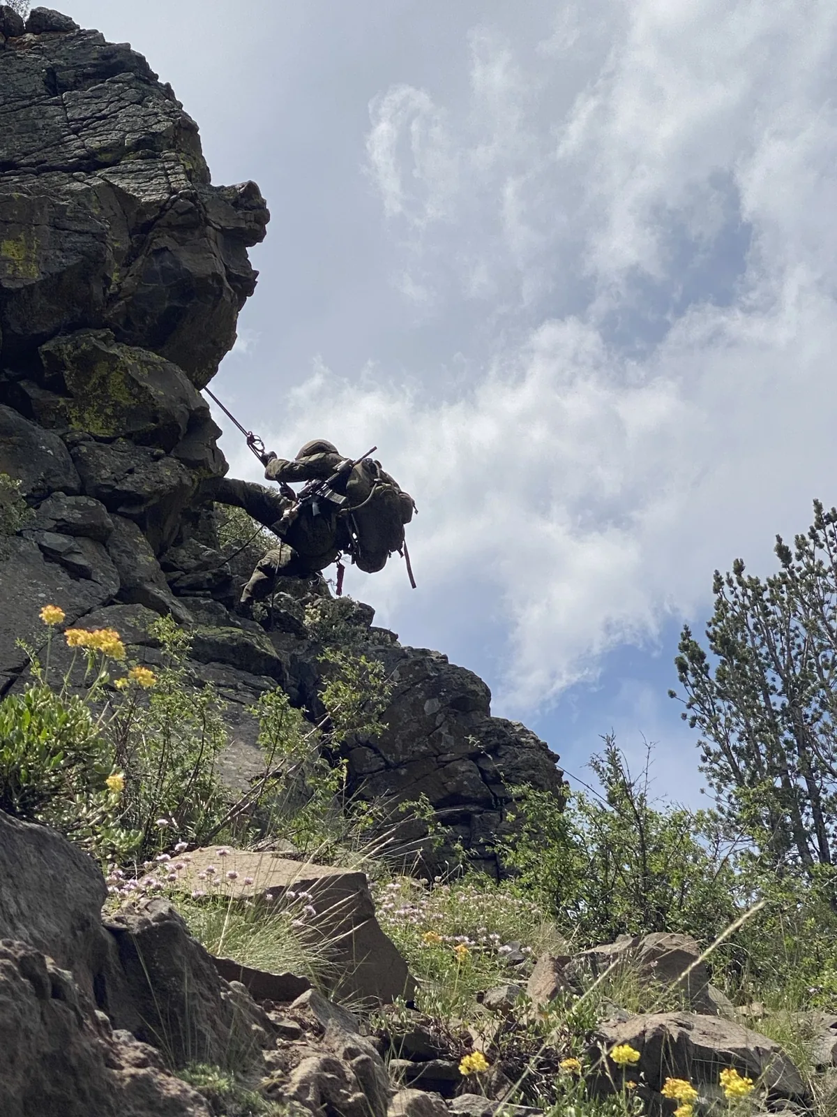 A Marine climbing a rocky cliff face under a full ruck in mountainous terrain