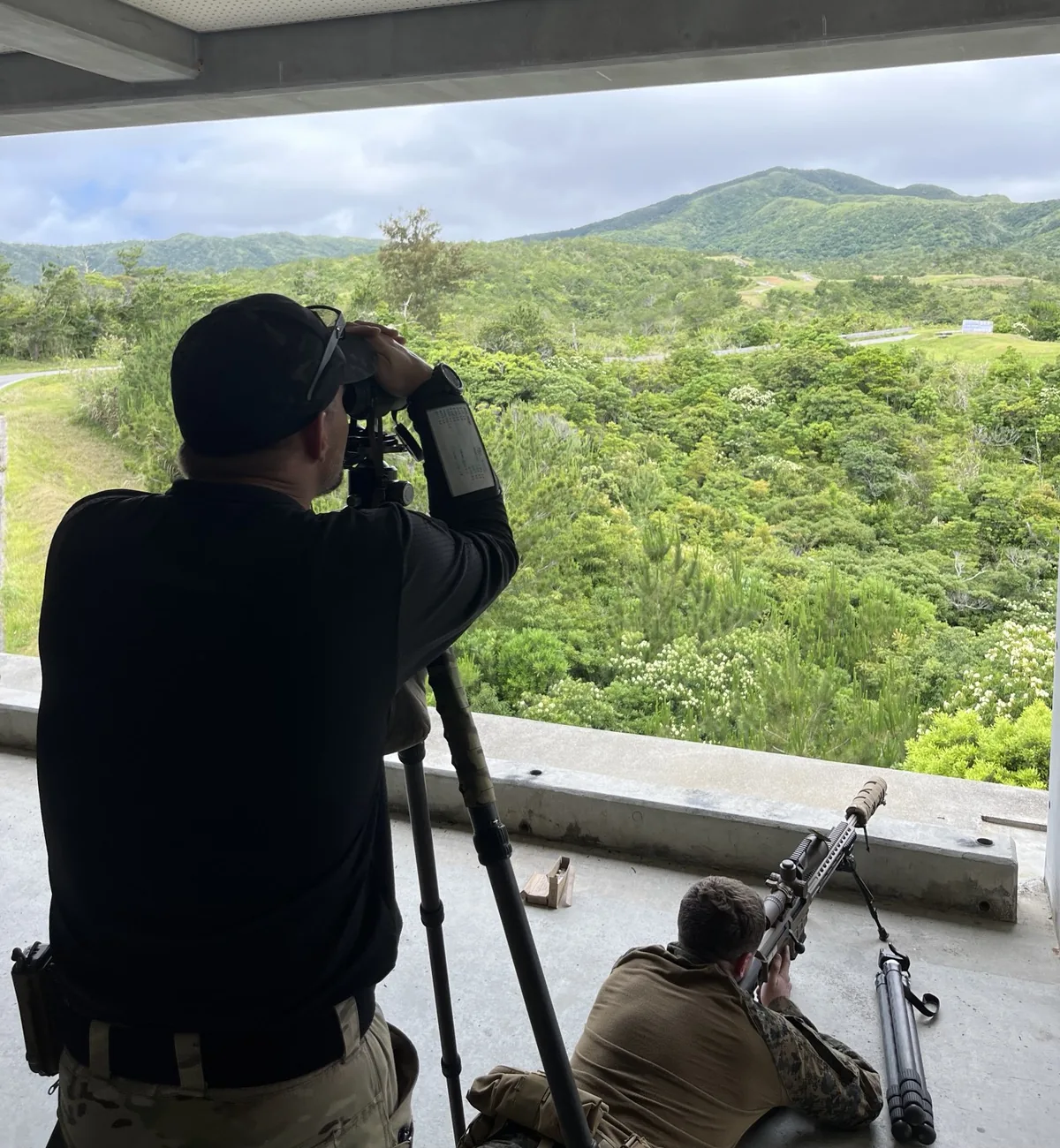 Eric Avvisato standing behind a tripod-mounted spotting scope while a shooter lies prone, green Okinawa hills in the background