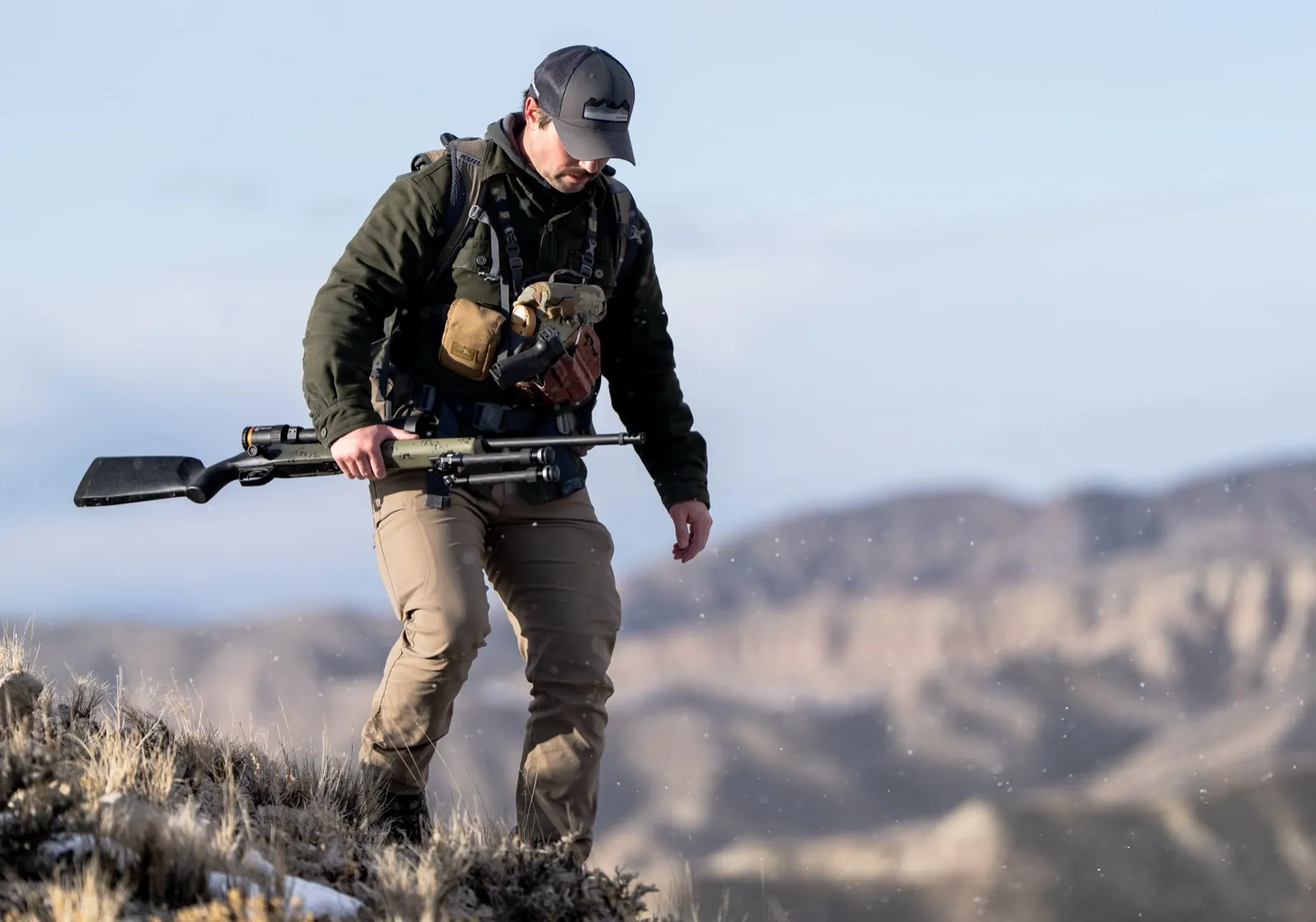 Ian Miner hiking a high ridge with his rifle carried across his chest, mountains behind him