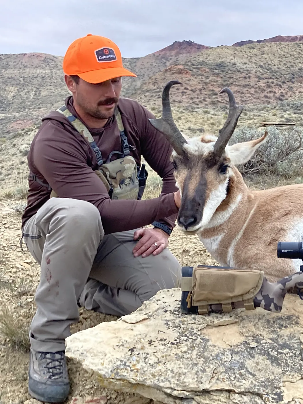 Ian Miner kneeling with a pronghorn buck he took, his rifle resting on a rock ledge