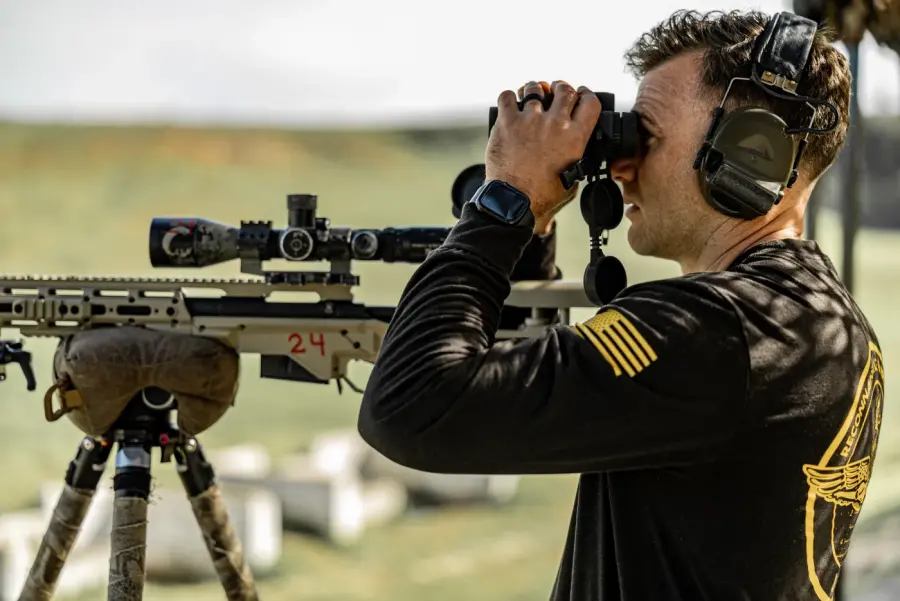 A Recon Sniper Course shooter glassing through binoculars beside his precision rifle