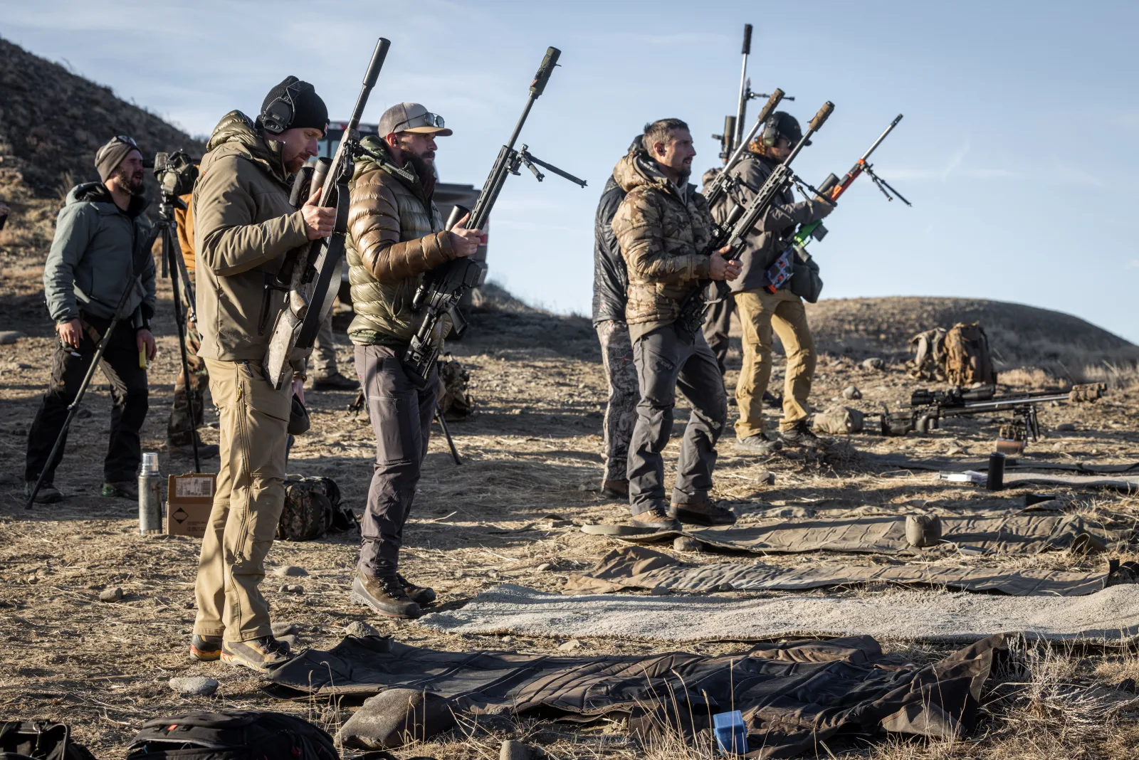 Shooters standing on the firing line holding their rifles upright, about to set them down onto mats — rifles not yet grounded.